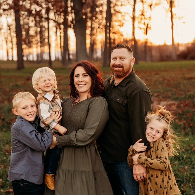 Family portrait featuring a mother, father, and three children smiling together in a forest during sunset. The family is dressed in casual, autumn attire, surrounded by trees with autumn foliage. This image captures a warm, joyful moment, perfect for family-themed content and seasonal celebrations.