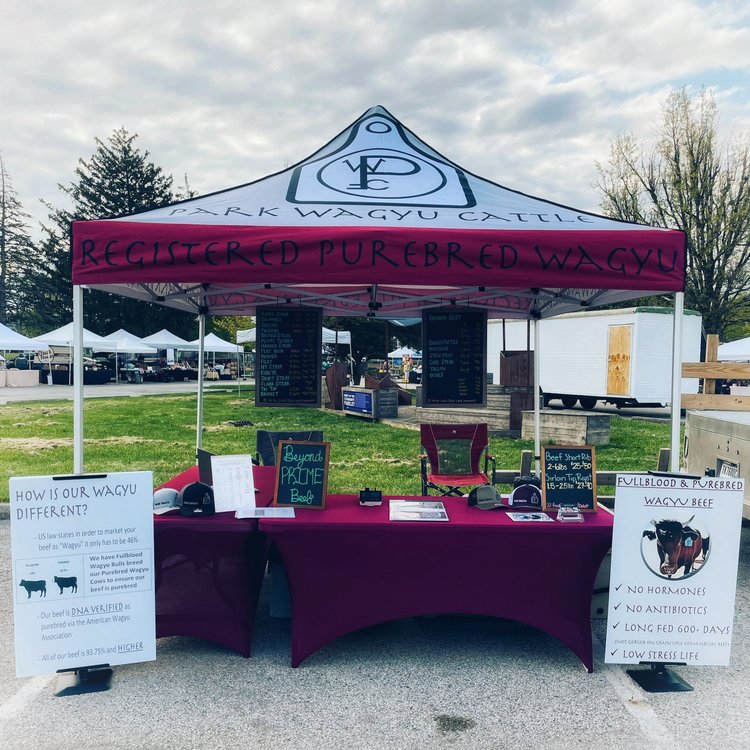 Wagyu beef market stall featuring a red canopy with "Registered Purebred Wagyu" signage, showcasing product information and benefits, including hormone-free and antibiotic-free options. The setup includes promotional materials and pricing for various cuts of Wagyu beef, set against a backdrop of a farmers' market environment.